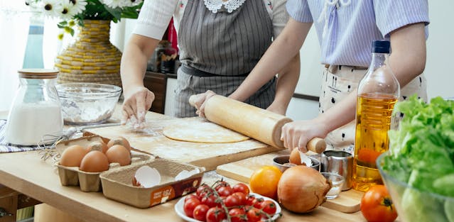 Close-up of people kneading dough in a pile of flour on a wooden table, with eggs, tomatoes, garlic, and pasta ingredients visible, suggesting the preparation of a meal.