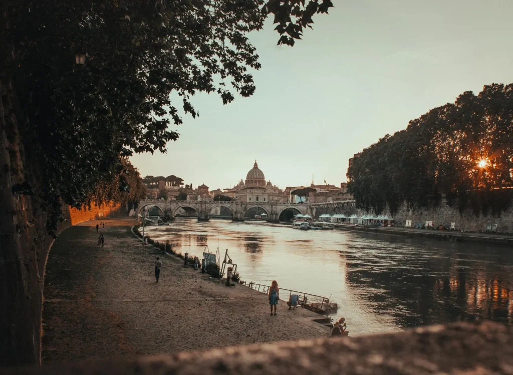 Scenic view of the Tiber River in Rome, Italy, with the dome of St. Peter's Basilica prominently visible in the background, bridges spanning the river, and the cityscape stretching along the banks, taken at dusk or dawn with warm lighting.