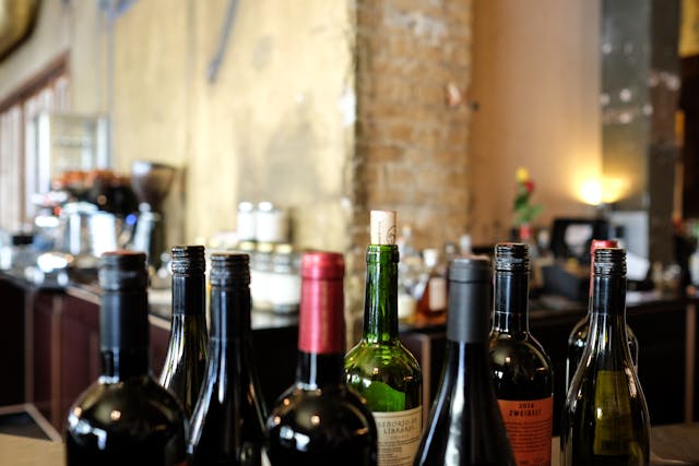 Close-up of several wine bottles of varying colors and sizes, some with corks and some with screw caps, arranged on a surface with a blurred restaurant or bar background featuring a coffee machine and other equipment