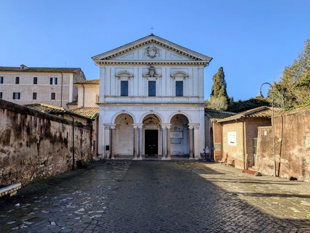 The facade of the Basilica di San Sebastiano fuori le mura in Rome, Italy, featuring a classical-style entrance with columns, a triangular pediment with an inscription, and a central doorway, with surrounding buildings and a cobblestone street visible under a clear blue sky.