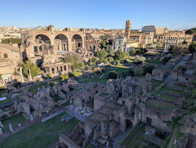 Panoramic view of the Roman Forum (Foro Romano) in Rome, Italy, showcasing the extensive ruins of ancient temples, basilicas, and other structures, with the Colosseum prominently visible in the background and the cityscape beyond.