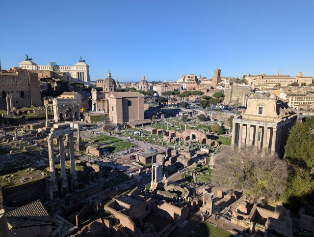 Panoramic aerial view of the Roman Forum in Rome, Italy, showcasing the extensive ruins of ancient temples, basilicas, and other structures, including the prominent Temple of Saturn with its remaining columns, the Arch of Titus, and the iconic Colosseum visible in the background, under a clear blue sky.