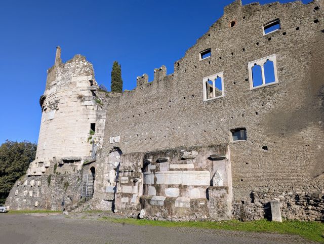 he Mausoleum of Caecilia Metella, an ancient Roman mausoleum with a distinctive cylindrical tower built of travertine, featuring remnants of decorative elements and inscriptions on the lower section, under a clear blue sky.