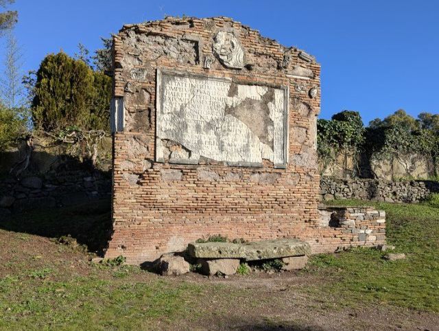 Ruined tomb on the ancient Appian Way (Via Appia Antica) in Italy, showing the remaining brick and stonework structure, a large rectangular inscription with weathered text, and the surrounding landscape with trees and grass.