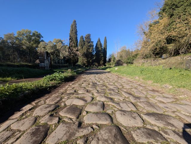 The ancient Appian Way (Via Appia Antica) in Italy, a historic road paved with cobblestones, stretching into the distance with tall trees lining both sides, under a clear blue sky.