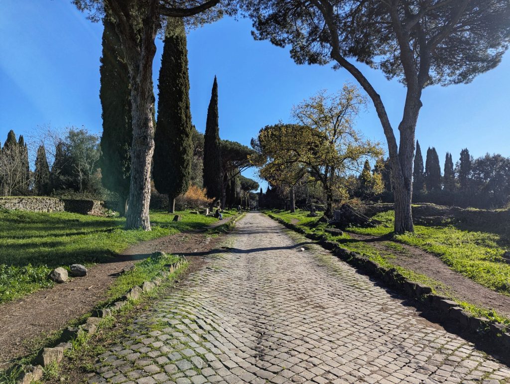 The ancient Appian Way (Via Appia Antica) in Italy, a historic road paved with cobblestones, stretching into the distance with tall trees lining both sides, under a clear blue sky.
