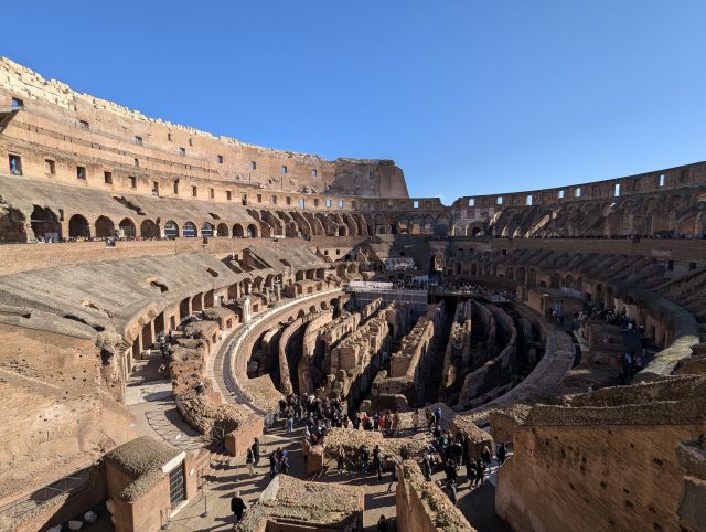 Interior view of the Colosseum, also known as the Flavian Amphitheatre, in Rome, Italy, showcasing the multiple levels of seating, the arena floor below, and the architectural details of the ancient Roman amphitheater, under a clear blue sky.