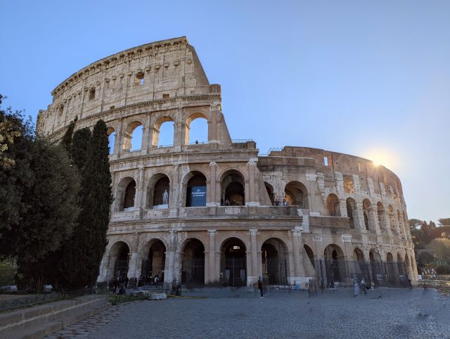 The Colosseum, also known as the Flavian Amphitheatre, in Rome, Italy, a large ancient Roman amphitheater with multiple levels of arches, under a blue sky with some clouds, with some trees visible on the left.