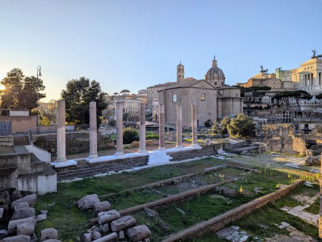 The ancient Roman Forum, Italy, featuring with prominent columns, the dome and facade of the Santi Luca e Martina church in the midground, and the Victor Emmanuel II monument ("Il Vittoriano") prominently featured in the distance, along with other ruins and structures under a clear blue sky.