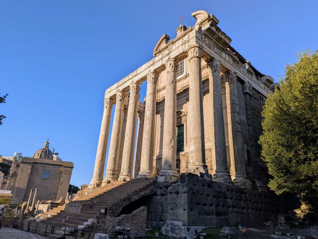 The Temple of Antoninus and Faustina, an ancient Roman temple in the Roman Forum, Rome, Italy, featuring a prominent portico with tall columns, a staircase leading up to the temple, and the ruins of other structures surrounding it, under a clear blue sky.