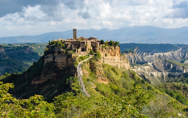 Aerial view of Civita di Bagnoregio, the "dying city," a medieval hilltop village in Italy, with a long pedestrian bridge spanning a deep gorge, surrounded by eroded cliffs and rolling hills.