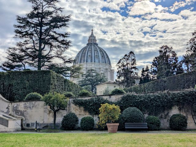 St. Peter's Basilica dome, a prominent architectural feature of Vatican City, seen from within the Vatican Gardens, with manicured hedges, trees, and a cloudy sky in the background.
