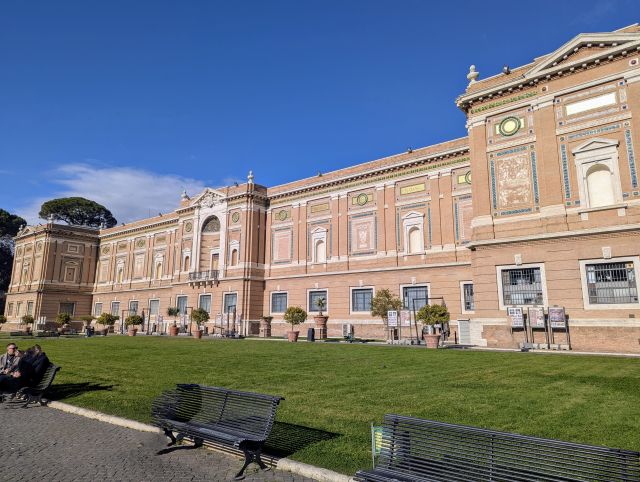 Facade of the Pinacoteca Vaticana within the Vatican Museums, featuring decorative architectural details, numerous windows, and a grassy area with benches in the foreground.