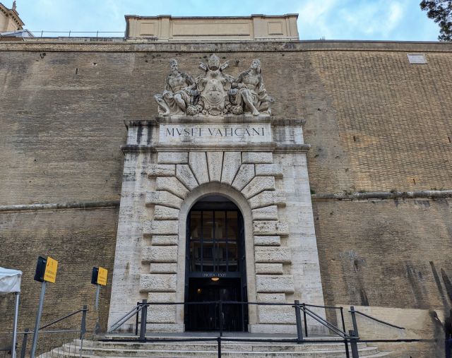Exterior view of the Vatican Museums, a stone structure with a large arched doorway, prominently displayed above, a coat of arms, and steps leading up to the entrance.