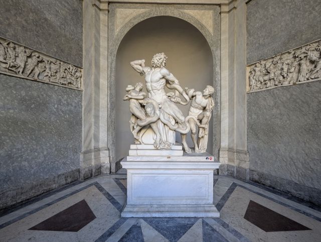 The Laocoön Group, a famous ancient Roman marble sculpture depicting Laocoön, a Trojan priest, and his two sons being attacked by sea serpents, displayed in the Vatican Museums, Rome, with other sculptures on the walls in the background.