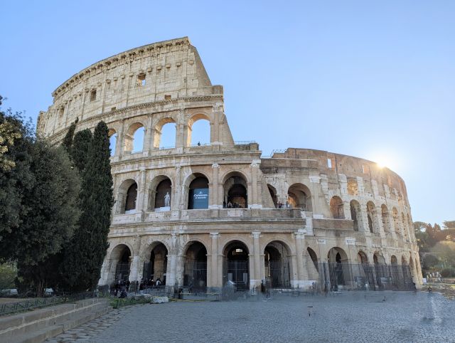The Colosseum, also known as the Flavian Amphitheatre, in Rome, Italy, a large ancient Roman amphitheater with multiple levels of arches, under a blue sky with some clouds, with some trees visible on the left.