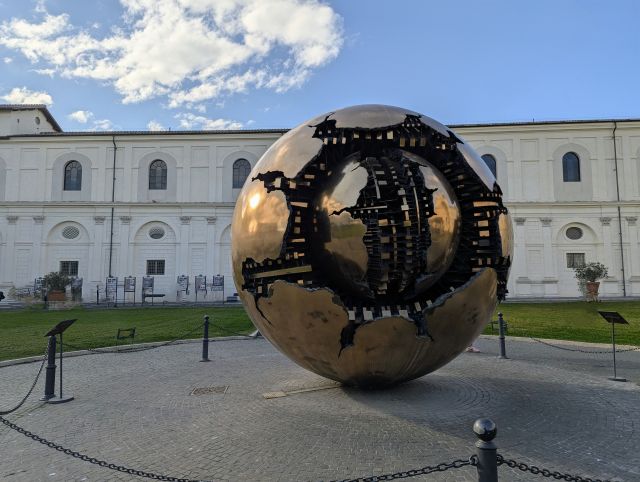 Sfera con Sfera" (Sphere Within Sphere), a large, golden bronze sculpture by Arnaldo Pomodoro, featuring a cracked outer sphere revealing a smaller sphere within, located in the Cortile della Pigna (Pine Cone Courtyard) of the Vatican Museums, Rome, with a light-colored building and blue sky in the background.
