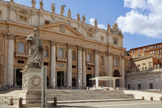 The grand facade of St. Peter's Basilica in Vatican City, featuring towering columns, numerous statues of saints atop the building and a wide staircase leading up to the entrance.