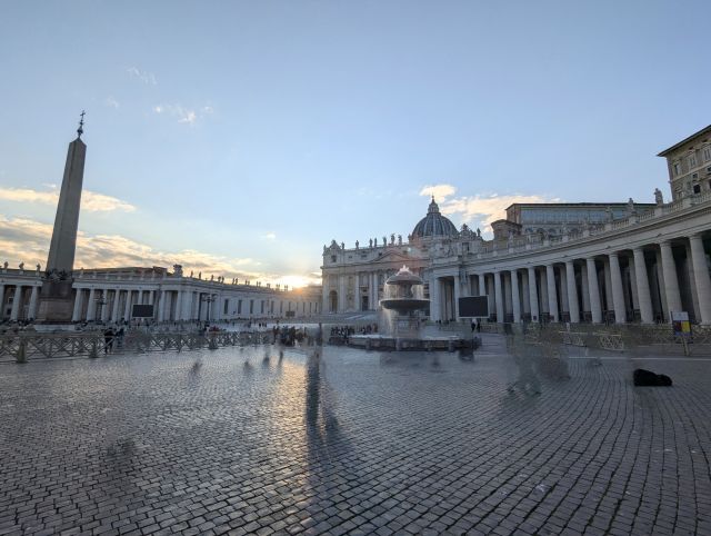 Saint Peter's Square and Basilica in Vatican City, a wide, paved square enclosed by colonnades, featuring an obelisk, fountains, and crowds of people, with the dome of the basilica visible against a bright sky at sunset.