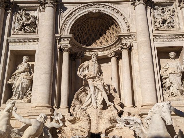 Close-up of the Trevi Fountain in Rome, Italy, showcasing the central sculptural group with Oceanus and his horses, intricate details of the Baroque architecture, cascading water, and tourists in the background.