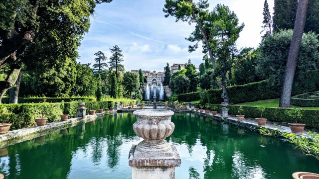 The intricate water gardens of Villa d'Este in Tivoli, Italy, featuring a long reflecting pool, numerous fountains, manicured hedges, and the villa on a hillside in the background.