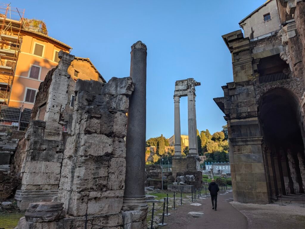 Udsigt over de antikke søjler i Apollon Sosianus-templet nær Marcellus-teateret i Rom, med en besøgende, der går gennem de historiske ruiner ved solnedgang.