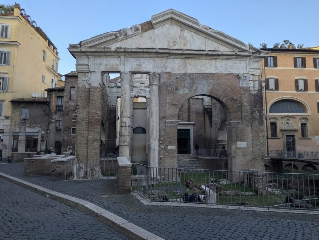 Frontudsigt over det antikke Portico d'Ottavia i Rom, der fremhæver dets historiske stensøjler, fronton og inskriptioner indrammet af omgivende bygninger.