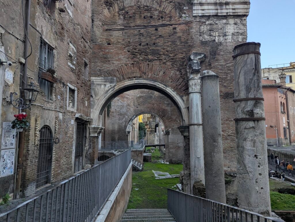 Udsigt gennem den gamle teglstensbuegang og søjler i Portico d'Ottavia i Rom, der afslører en gadescene og trapper, der fører ned til det arkæologiske område.