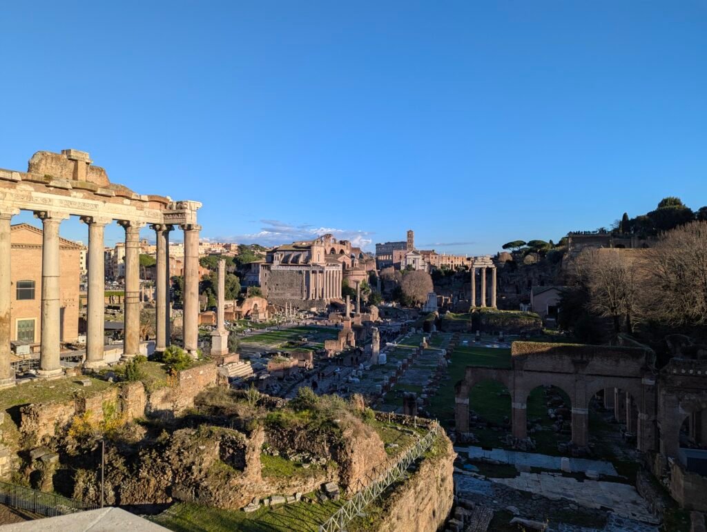 Wide panoramic view over the Roman Forum ruins with ancient columns in the foreground and the Colosseum in the distance