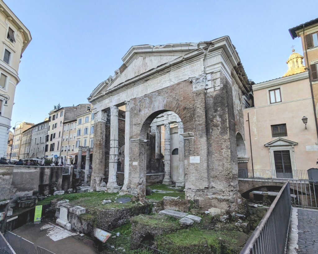 Utsikt over de antikke ruinene av Portico d'Ottavia i Roma, med sin historiske teglsteinsarkitektur, søyler og omliggende bygninger under en klar blå himmel.