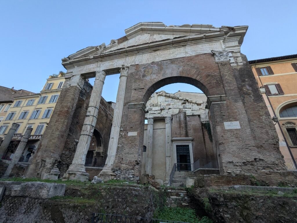 Lavvinkelutsikt over de antikke ruinene og den monumentale inngangen til Portico d'Ottavia i Roma, som viser dens historiske teglsteinsbuer, søyler og inskripsjoner mot en blå himmel.