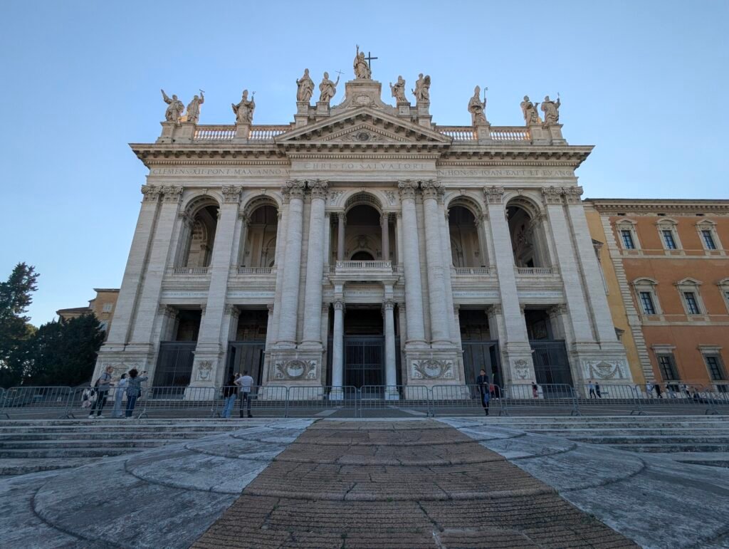 LFrontutsikt over den storslåtte og monumentale fasaden til San Giovanni in Laterano-basilikaen i Roma, med ruvende søyler, store buede innganger og statuer langs taklinjen mot en klar blå himmel.