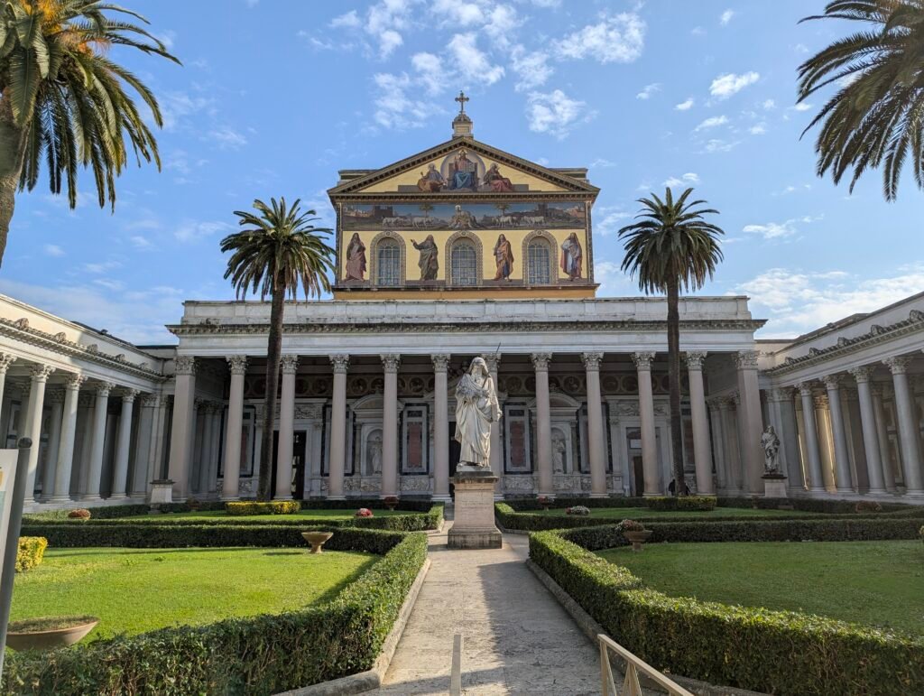 Utsikt over den storslåtte gårdsplassen, søylegangen, palmettrærne, statuen av Paulus og den mosaikkdekorerte fasaden til basilikaen San Paolo fuori le mura i Roma under en klar blå himmel.