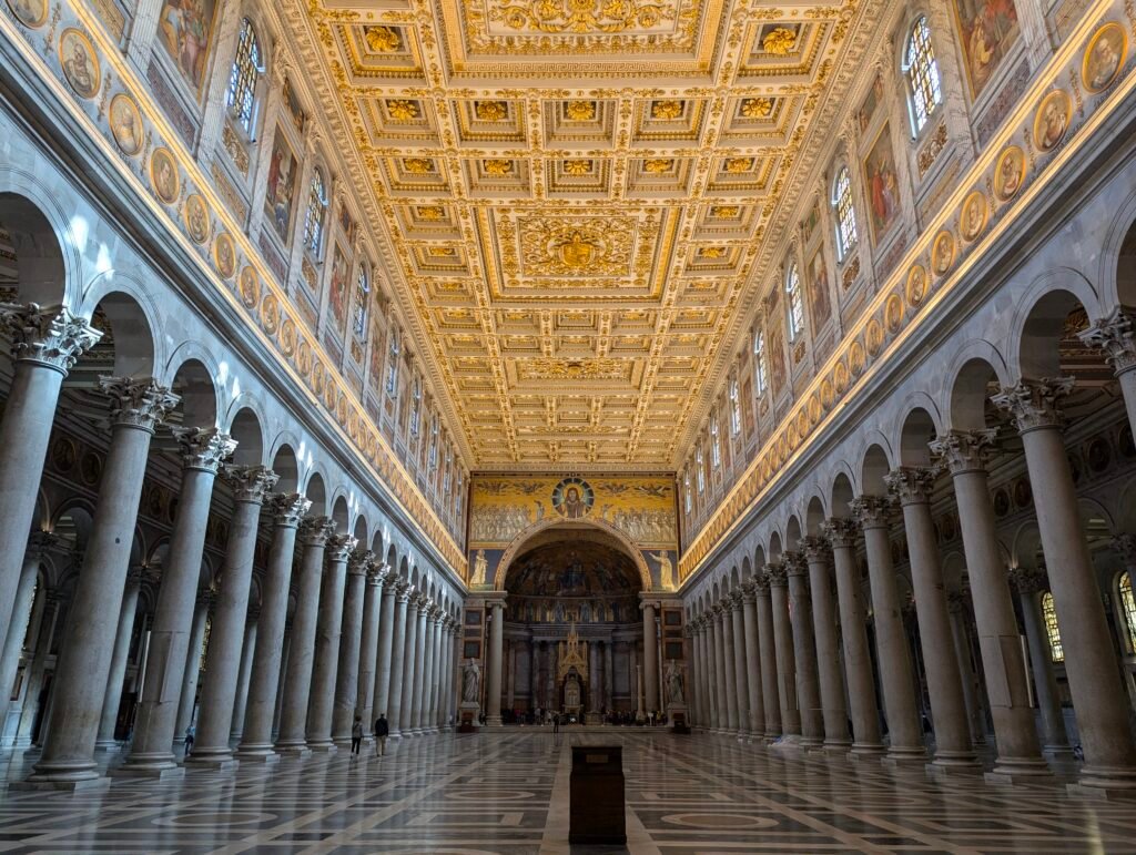 Innvendig utsikt over det storslåtte hovedskipet i basilikaen San Paolo fuori le mura i Roma, med endeløse rader av monolittiske søyler, et storslått forgylt kassetttak og apsismosaikken i det fjerne.