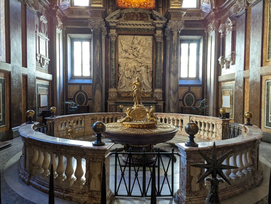 Innvendig utsikt over det utsmykkede døpefontkapellet inne i den pavelige basilikaen Santa Maria Maggiore i Roma, med en gullfarget døpefont, marmorbalustrade og klassiske relieffskulpturer.