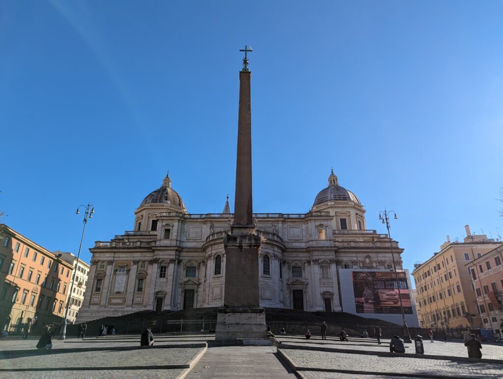 Utsikt over den majestetiske bakre fasaden og kuplene til den pavelige basilikaen Santa Maria Maggiore i Roma, med den høye Esquiline-obelisken i midten av den solrike plassen.