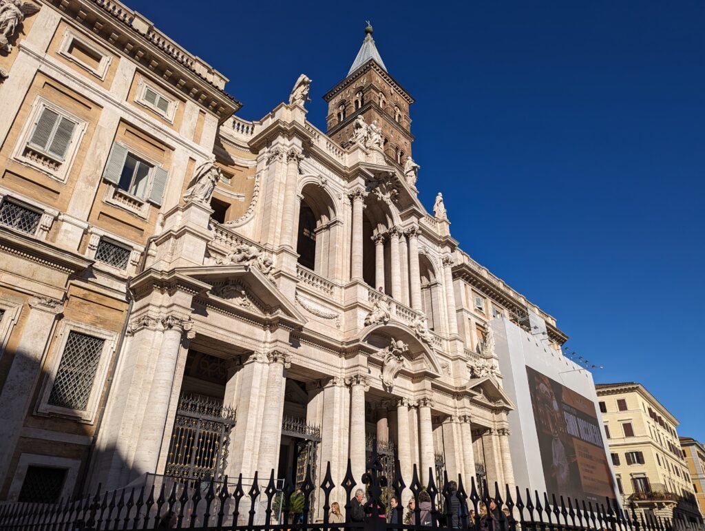 Lavvinkel utvendig utsikt over den storslåtte fasaden og inngangen til den pavelige basilikaen Santa Maria Maggiore i Roma, med besøkende som går og samles på den solrike plassen under en klar blå himmel.