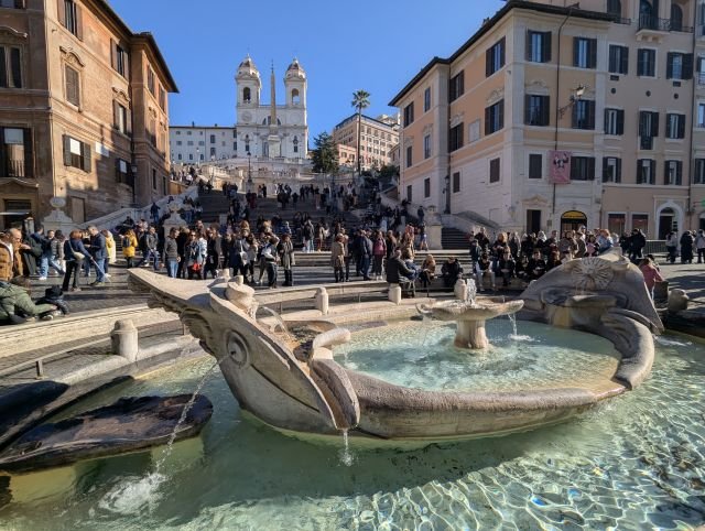 Utsikt over Spanske trappen og Barcaccia-fontenen på Piazza di Spagna i Roma, med besøkende som nyter den solfylte dagen.
