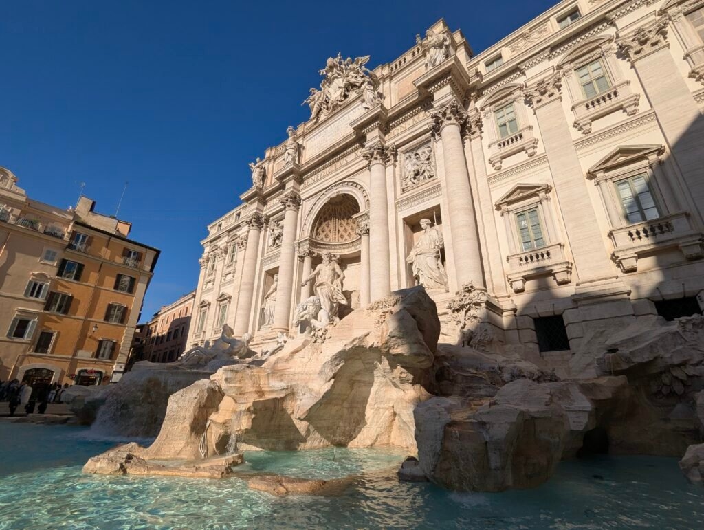 Imponerende lavvinkelutsikt over Trevifontenen i Roma med sentralstatuen av Oceanus og barokkfasaden mot en klar blå himmel.