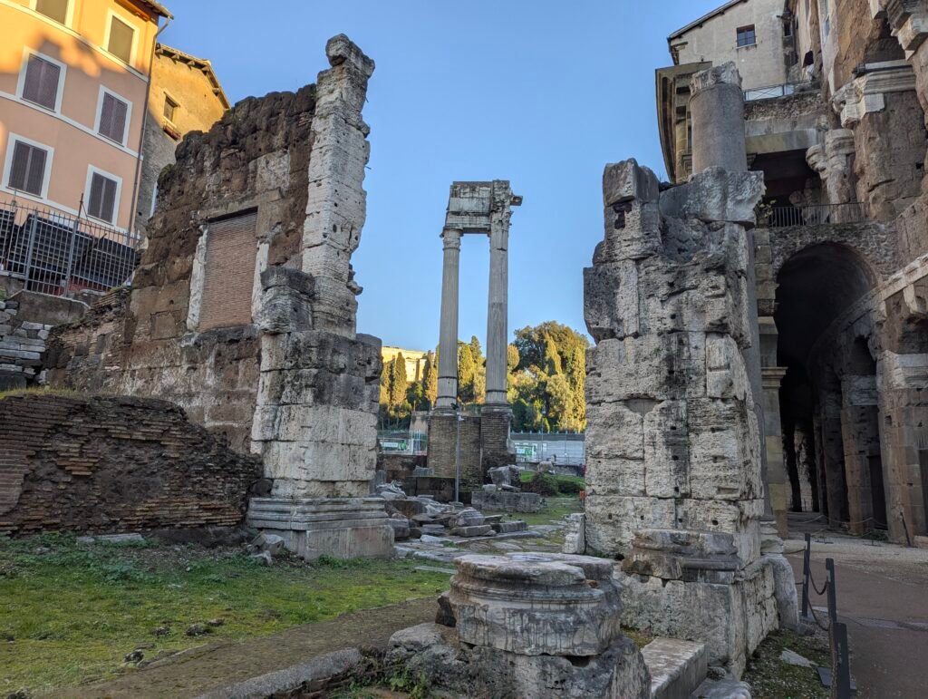Vy som ramar in de stående pelarna i Apollon Sosianus-templet genom gamla stenruiner i Rom, med en modern byggnad och klar blå himmel i bakgrunden.