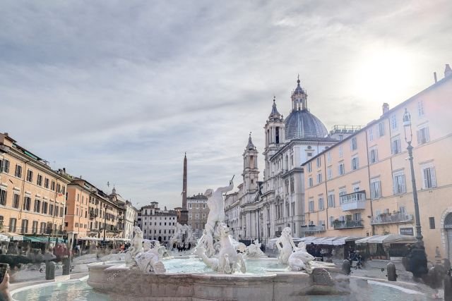Utsikt över Neptunusfontänen på Piazza Navona i Rom, med omgivande historiska byggnader och kyrkokupol under en ljus himmel.