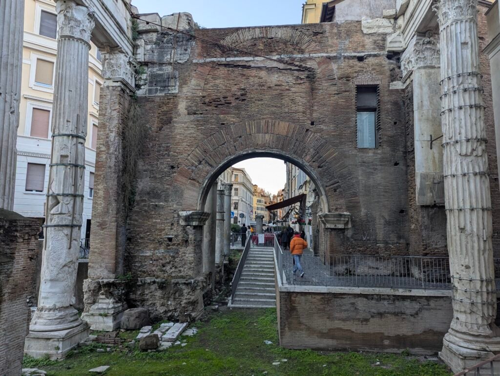 Vy genom den gamla tegelvalvsporten och pelarna i Portico d'Ottavia i Rom, som avslöjar en gatuscen och trappor som leder ner till den arkeologiska platsen.
