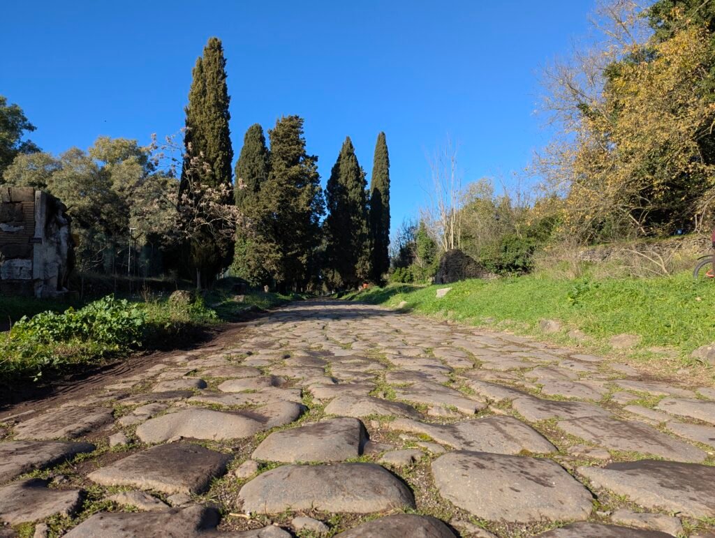 Lågvinkelperspektiv av den gamla romerska gatstensvägen på Via Appia Antica, flankerad av höga cypresser och frodig landsbygd under en blå himmel.